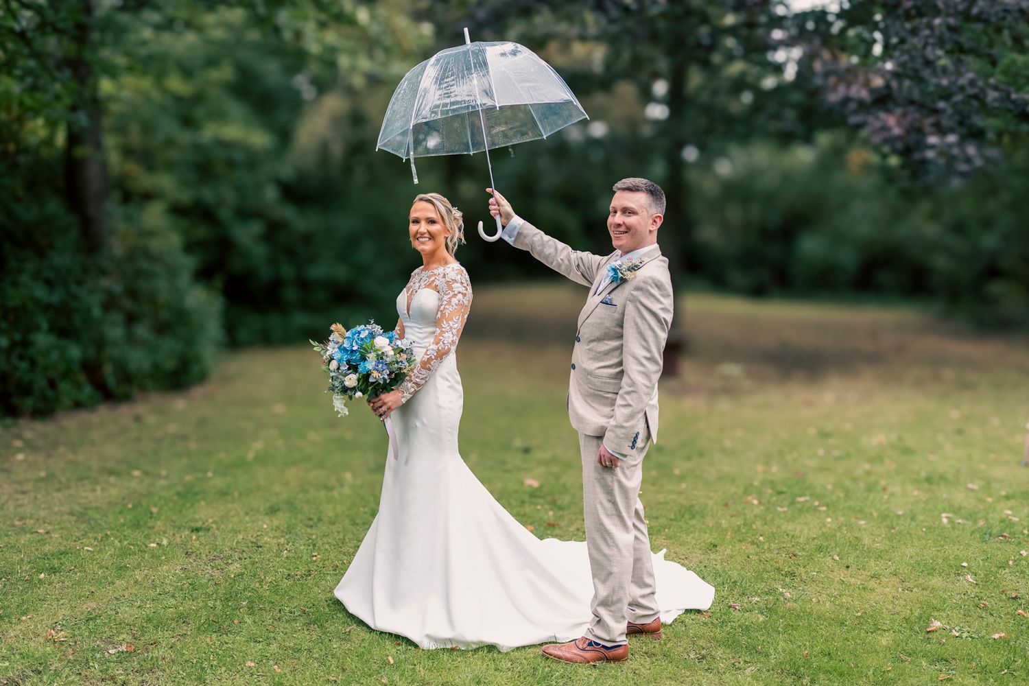 Bride in white lace gown holding blue bouquet stands with groom in tan suit holding clear umbrella on green lawn.
