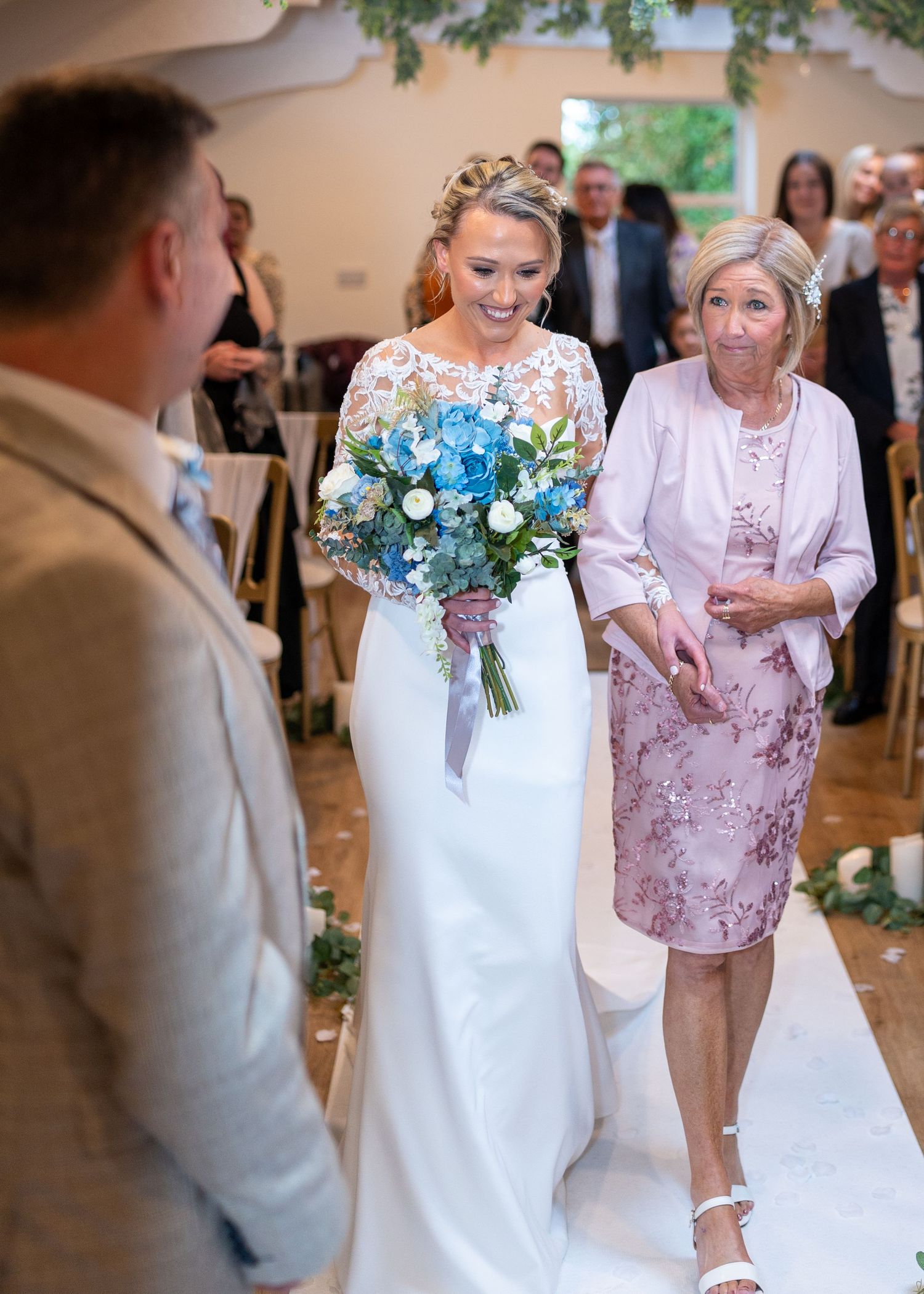 Bride in white lace gown holding blue bouquet walks down aisle with older woman at elegant wedding ceremony. Dave Martin Photography