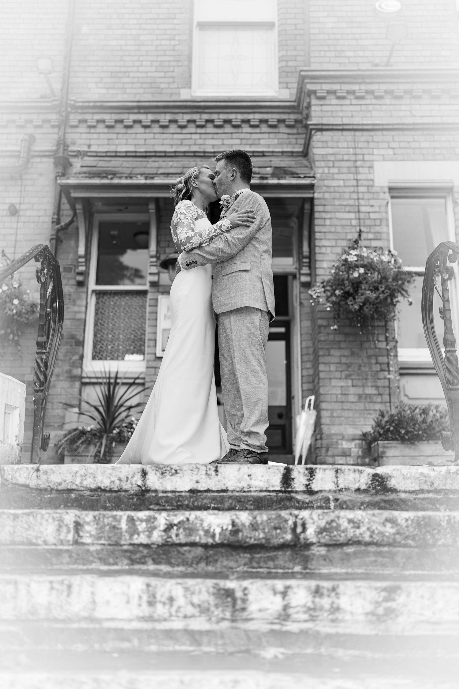 Black and white photo of a bride and groom embracing on stone steps outside a historic brick building.