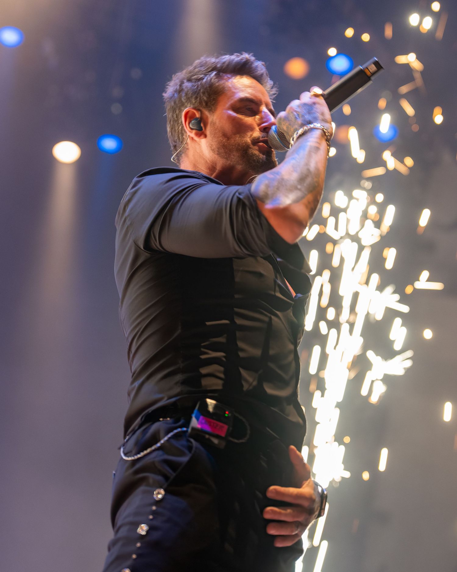Male singer in black outfit performs passionately with golden sparks exploding behind him on stage. Dave Martin celebrity Photography