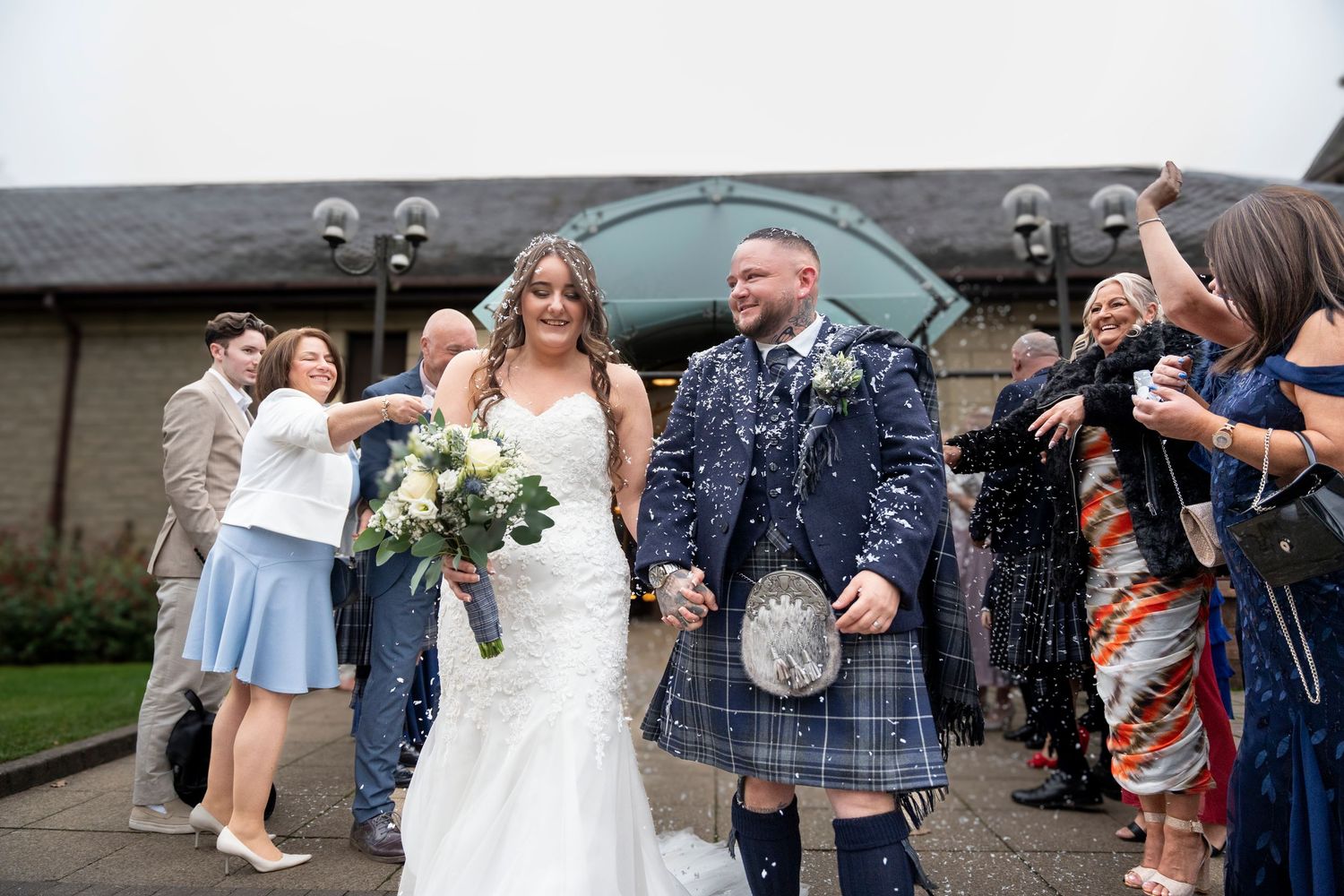 Bride and groom exit wedding venue in Scottish kilt as guests shower them with confetti. Dave Martin Photographer Northern Ireland