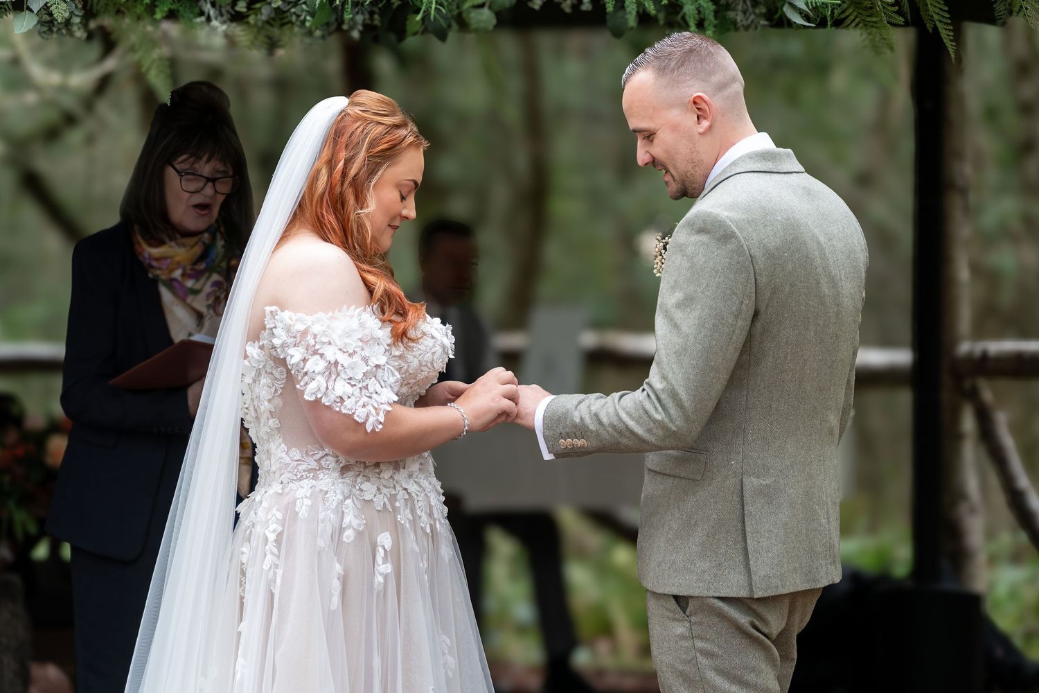 Redhead bride in floral gown exchanges rings with groom during outdoor woodland wedding ceremony. Dave Martin Photographer Northern Ireland