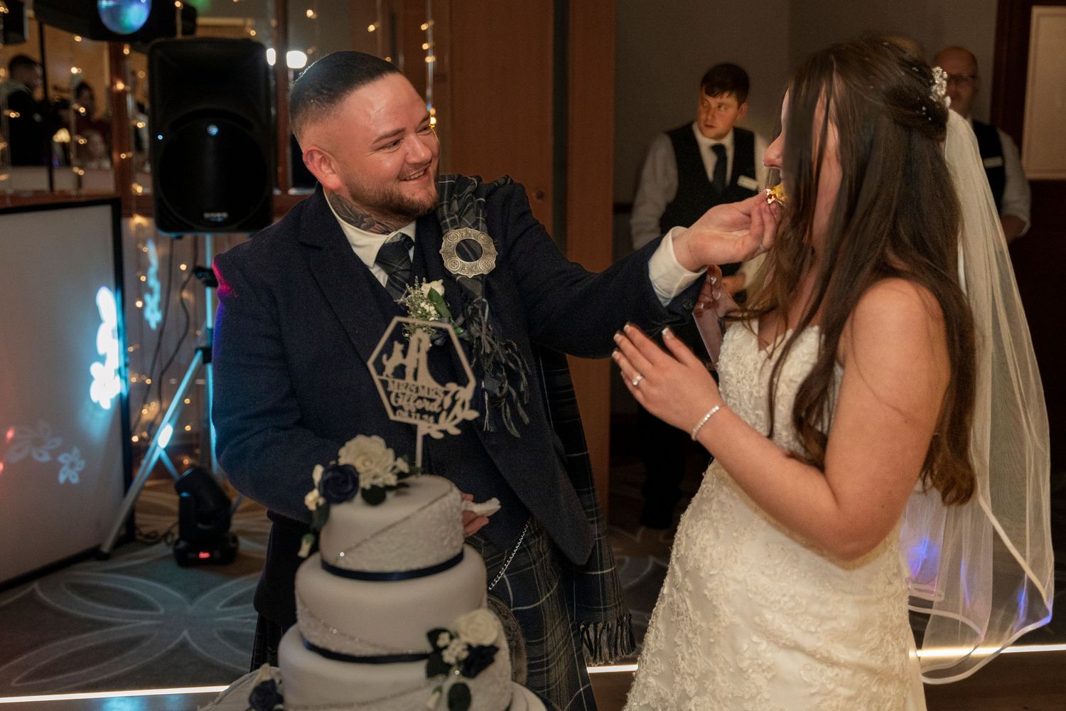 Groom in blue Scottish kilt feeding wedding cake to bride at reception celebration. Dave Martin Photographer Northern Ireland