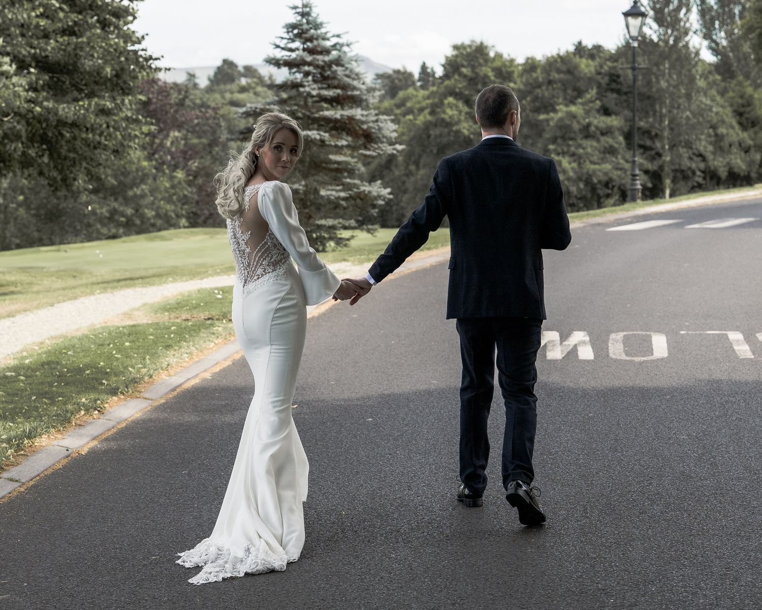 Bride in white lace gown holds hands with groom in navy suit walking on road surrounded by greenery.   ni wedding photographer Dave Martin