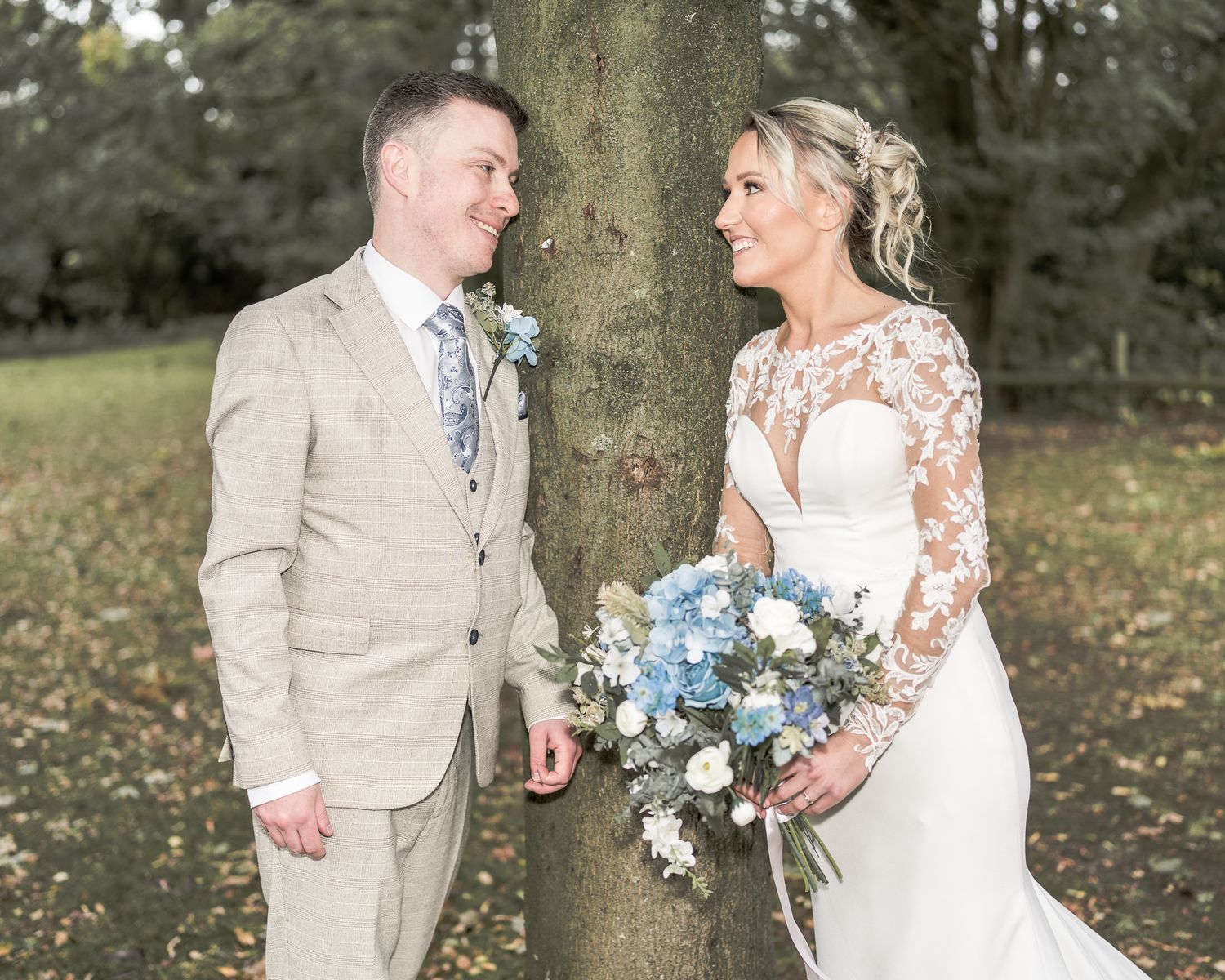 Bride and groom stand by a tree, she holds blue and white bouquet, he wears tan suit with floral tie, outdoors. Dave Martin wedding Photos