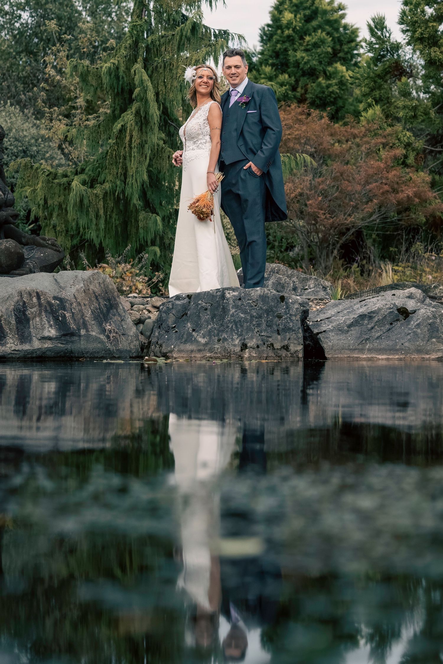 Bride and groom standing on rocks by a tranquil pond, their reflection mirrored in the still water. Northern ireland photographer