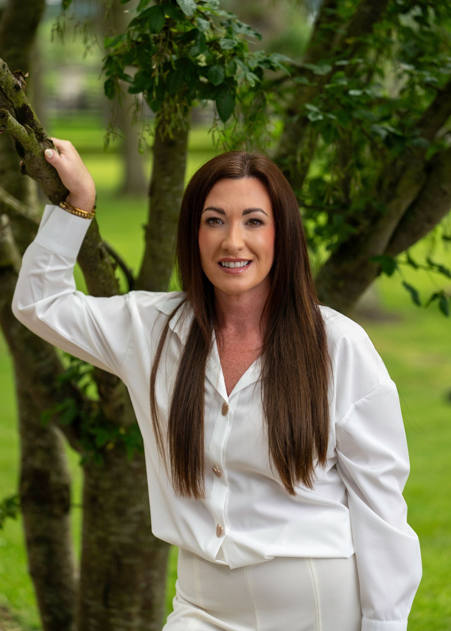 Woman with long dark hair wearing a white outfit posing outdoors beside a tree in a lush green garden. Dave Martin Photography