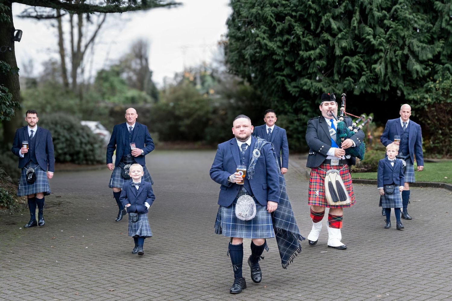 Scottish wedding party in blue kilts led by bagpiper walking through hotel grounds. Dave Martin Photographer Northern Ireland