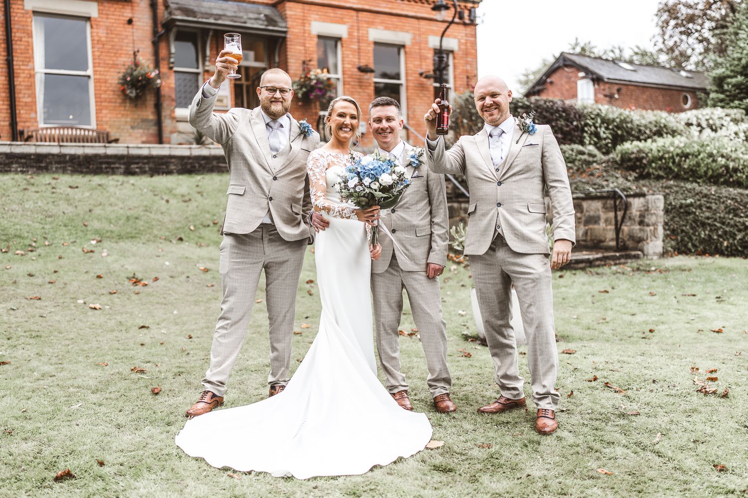 Bride in white gown poses with three groomsmen in tan suits on a green lawn outside a brick building. Dave Martin wedding Photographer NI