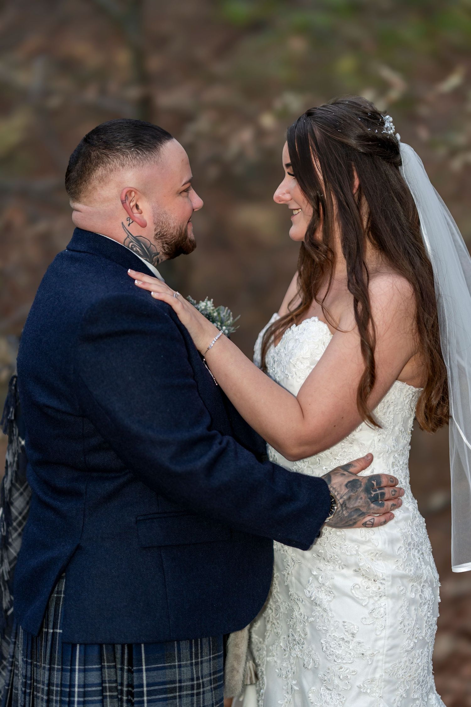 Smiling bride and tattooed groom in blue kilt share tender moment outdoors. Dave Martin Photographer Northern Ireland