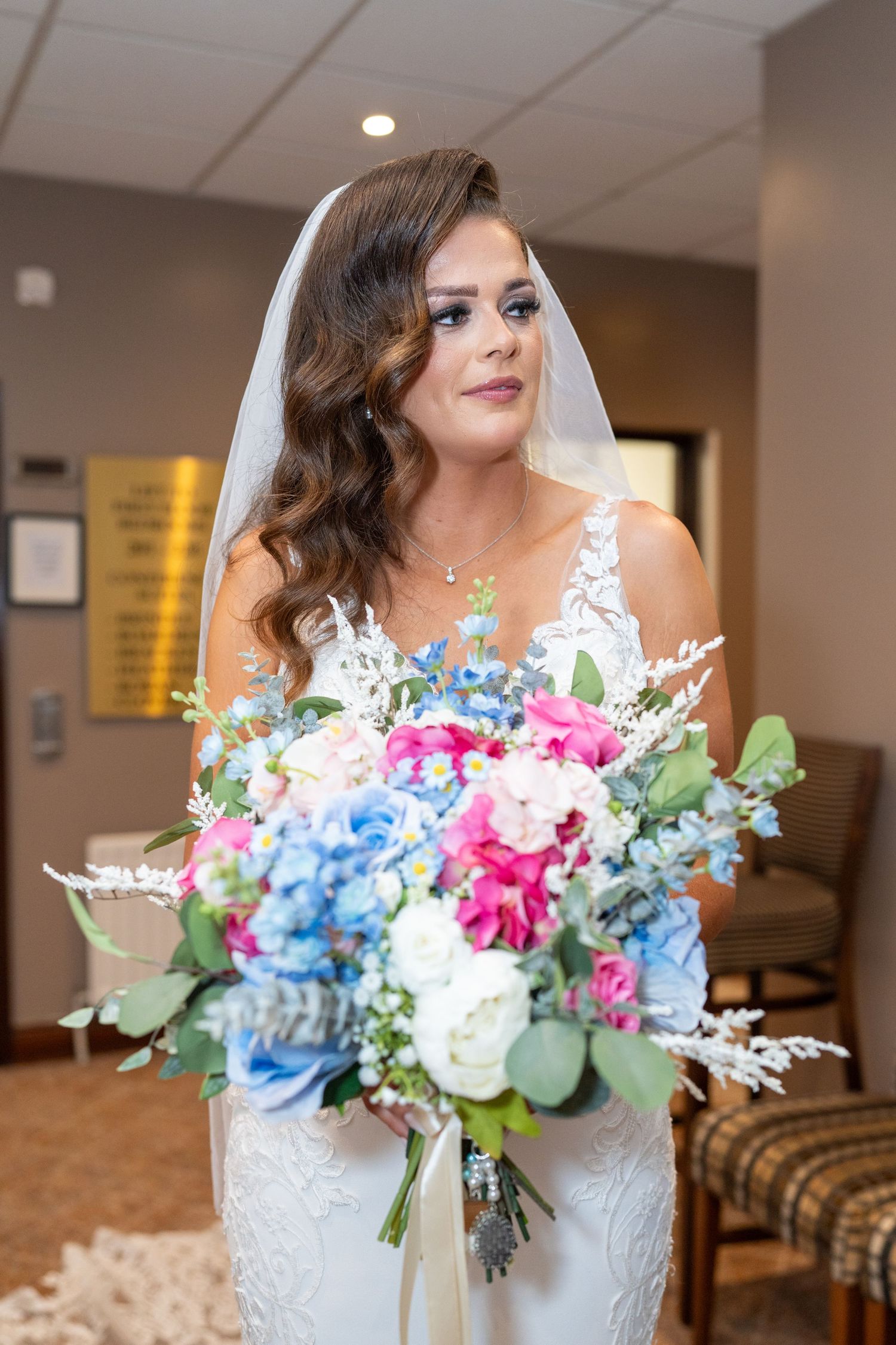 Bride in lace gown holds a colorful bouquet of pink, blue, and white flowers indoors. Dave Martin Photographer Northern Ireland