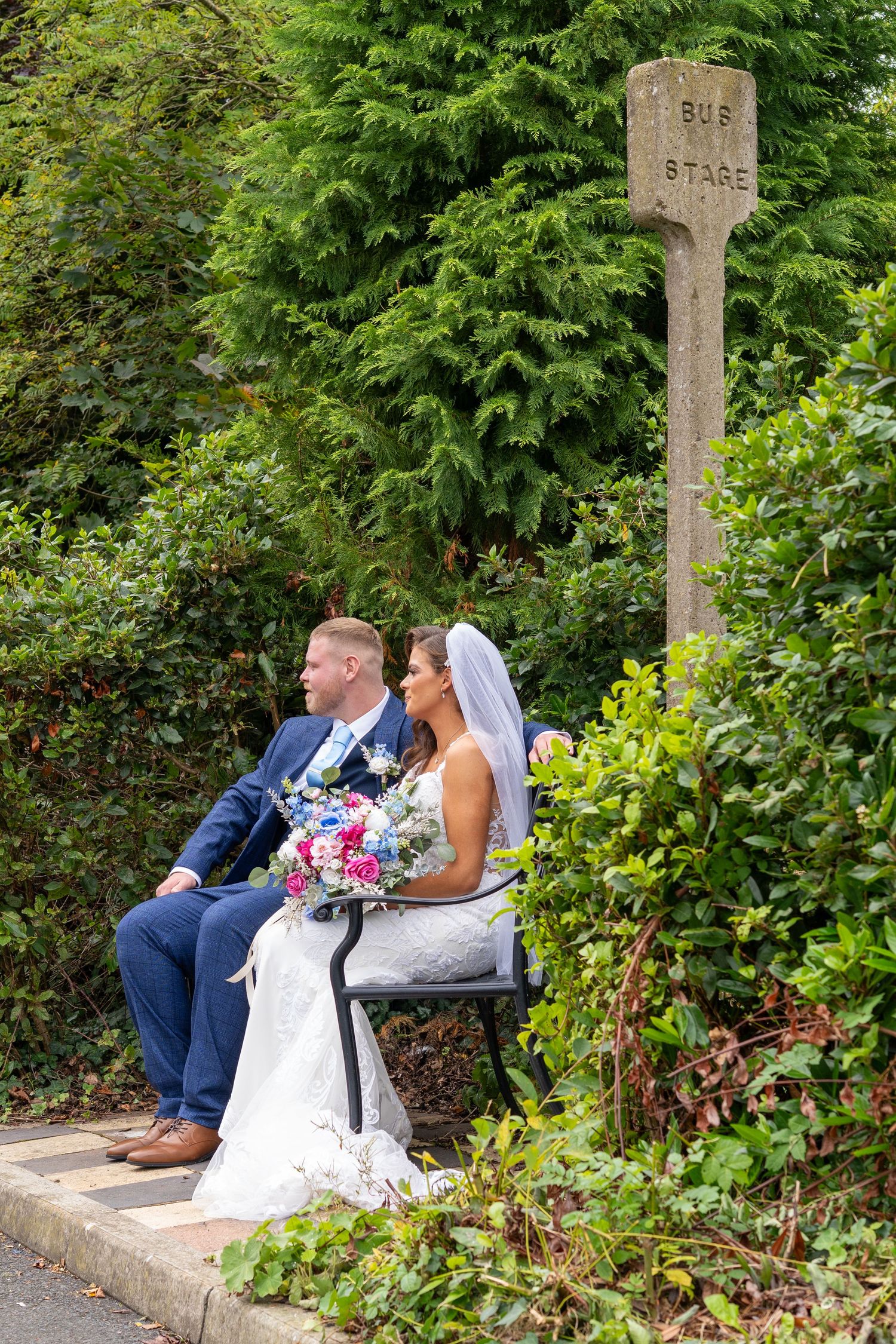 Bride and groom sit together at a vintage bus stop sign surrounded by lush greenery. Dave Martin Photographer Northern Ireland