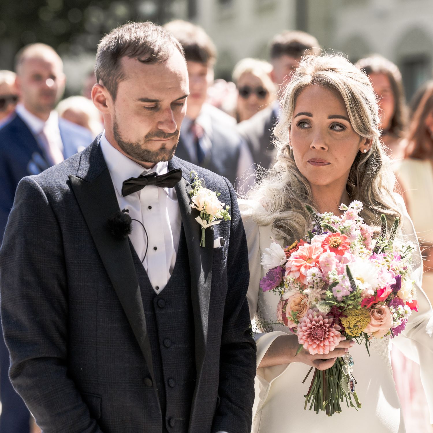 Bride holding pink floral bouquet stands beside groom in black tuxedo at outdoor wedding ceremony. Dave Martin wedding Photographer NI