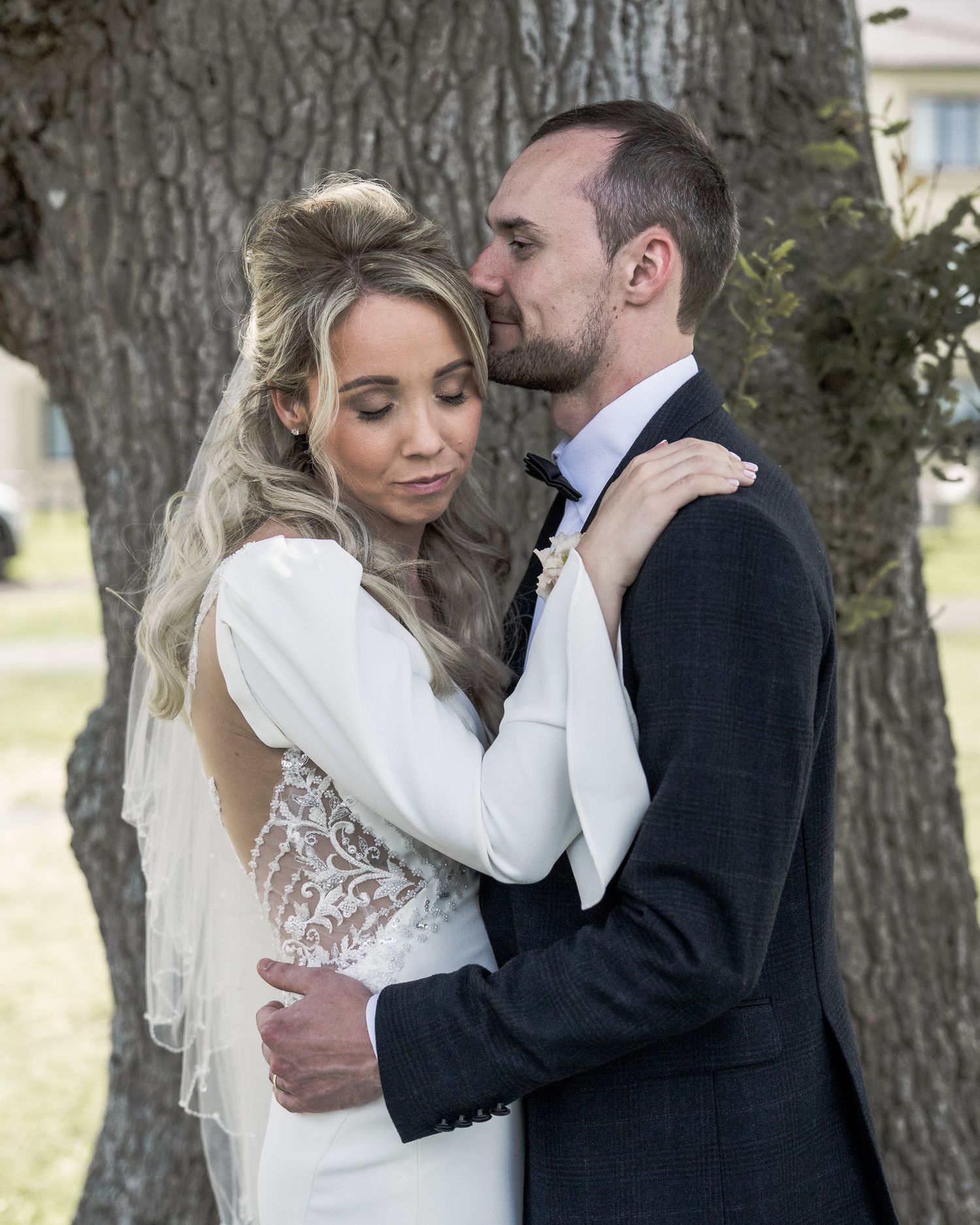 A bride and groom share an intimate embrace near a large tree, dressed in wedding attire outdoors. Northern ireland weddings