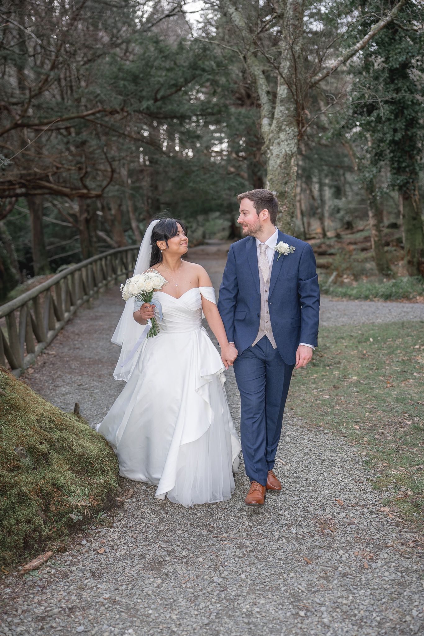 A bride and groom walk hand-in-hand along a woodland path, she in a white gown with veil, he in a navy suit. Dave Martin wedding Photograph
