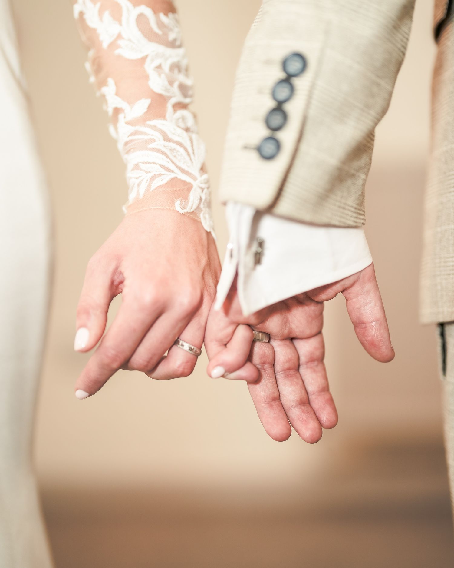 Bride and groom holding hands, showcasing wedding rings, lace bridal sleeve and beige suit at their wedding ceremony. Dave Martin wedding