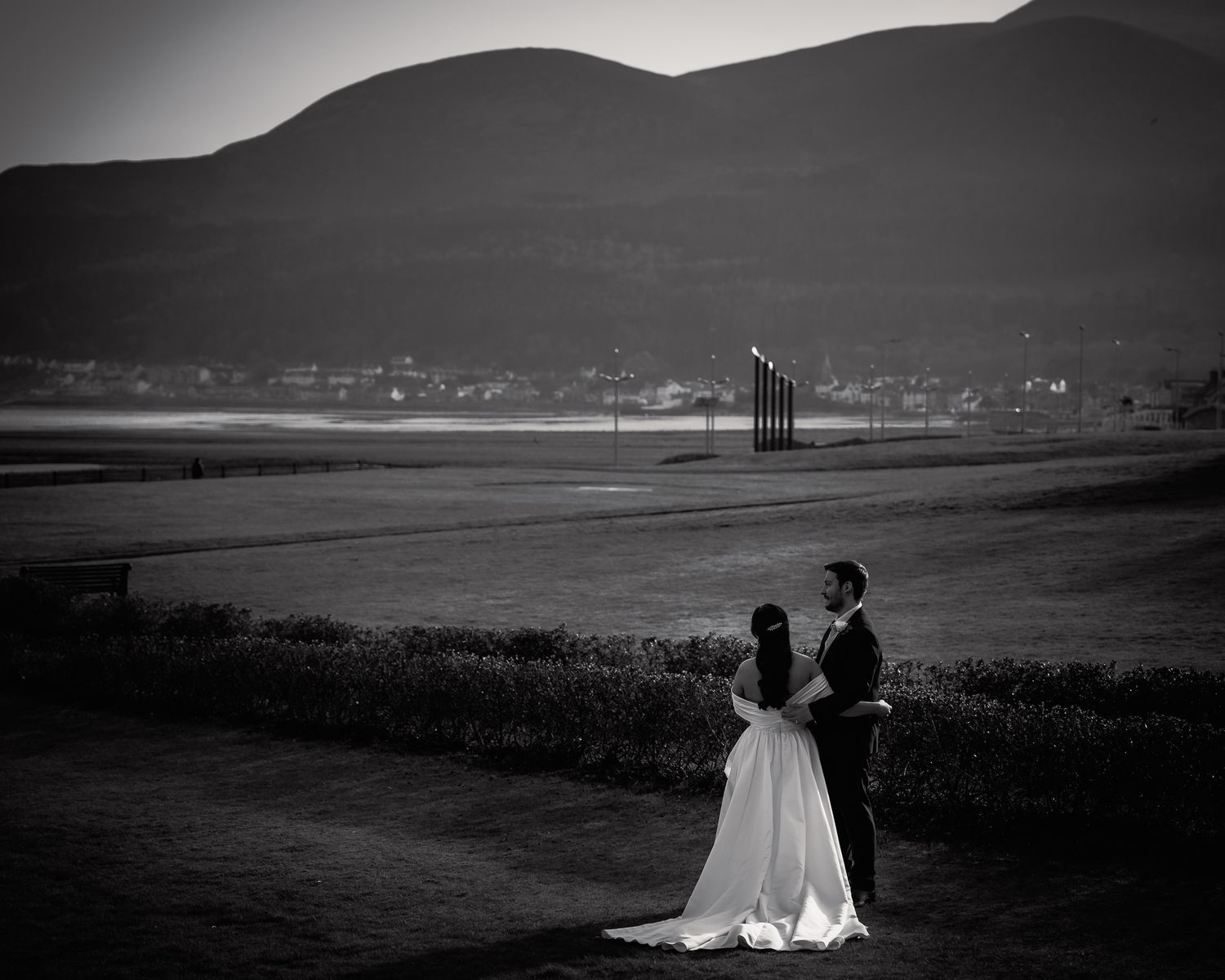 Black and white wedding photo of bride and groom standing on a path with mountains and water in the background.