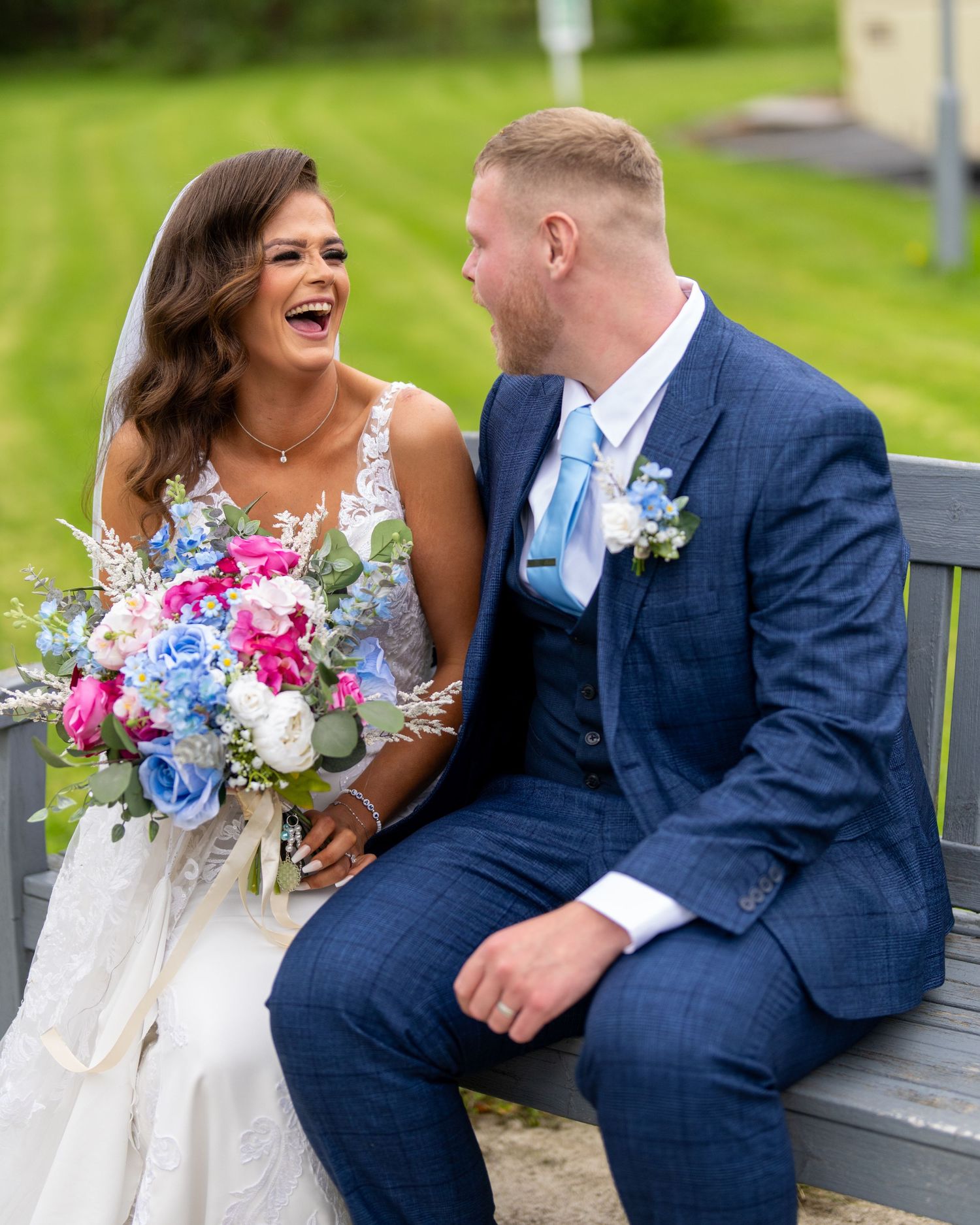 Bride and groom laughing joyfully on a bench, bride holding colorful blue and pink bouquet. Dave Martin Photographer Northern Ireland