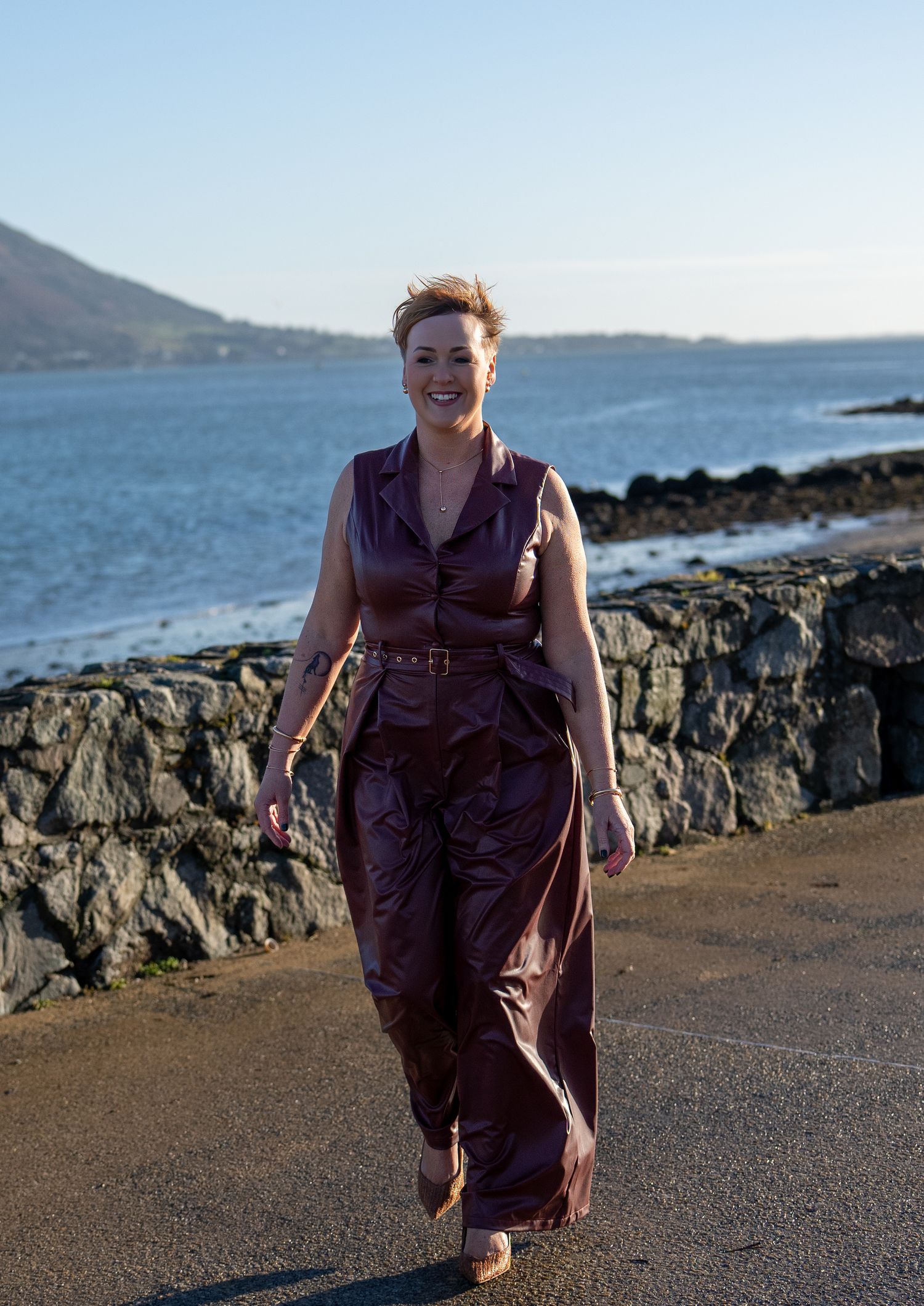 Woman in purple jumpsuit walking along coastal road with sea and mountains behind. Dave Martin Photography Northern Ireland
