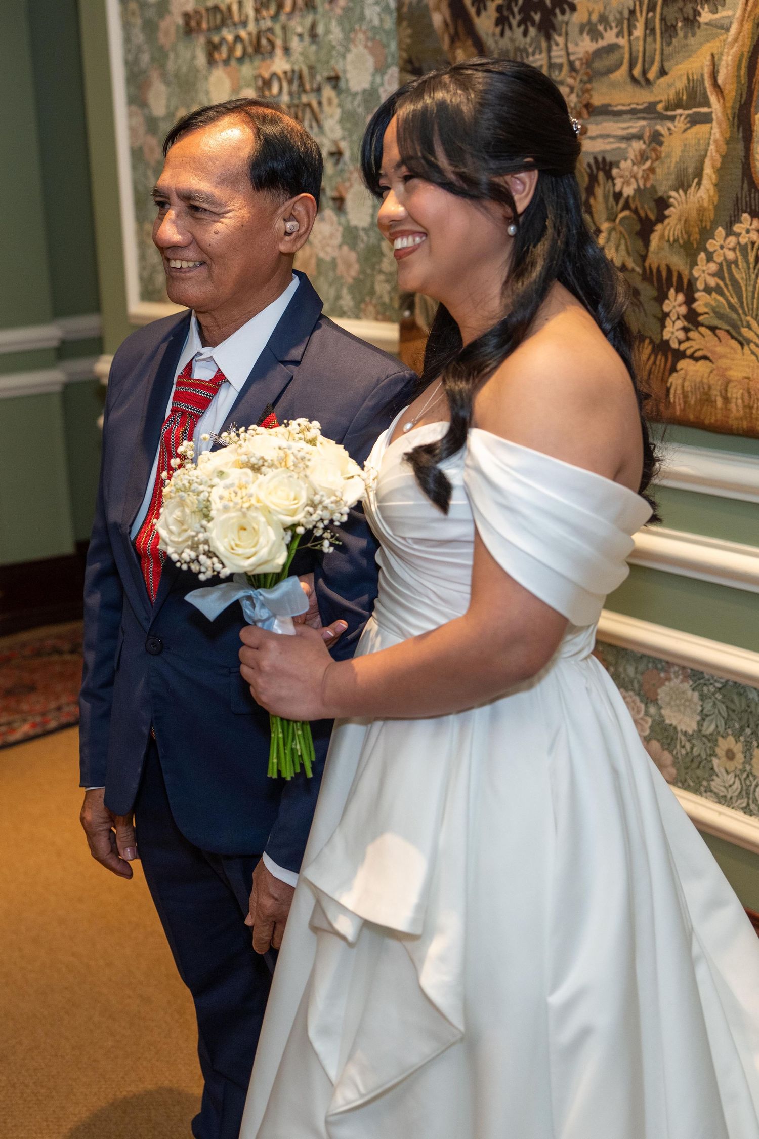 Bride in white off-shoulder gown holds white rose bouquet while walking with older man in navy suit. Slieve Donard Hotel