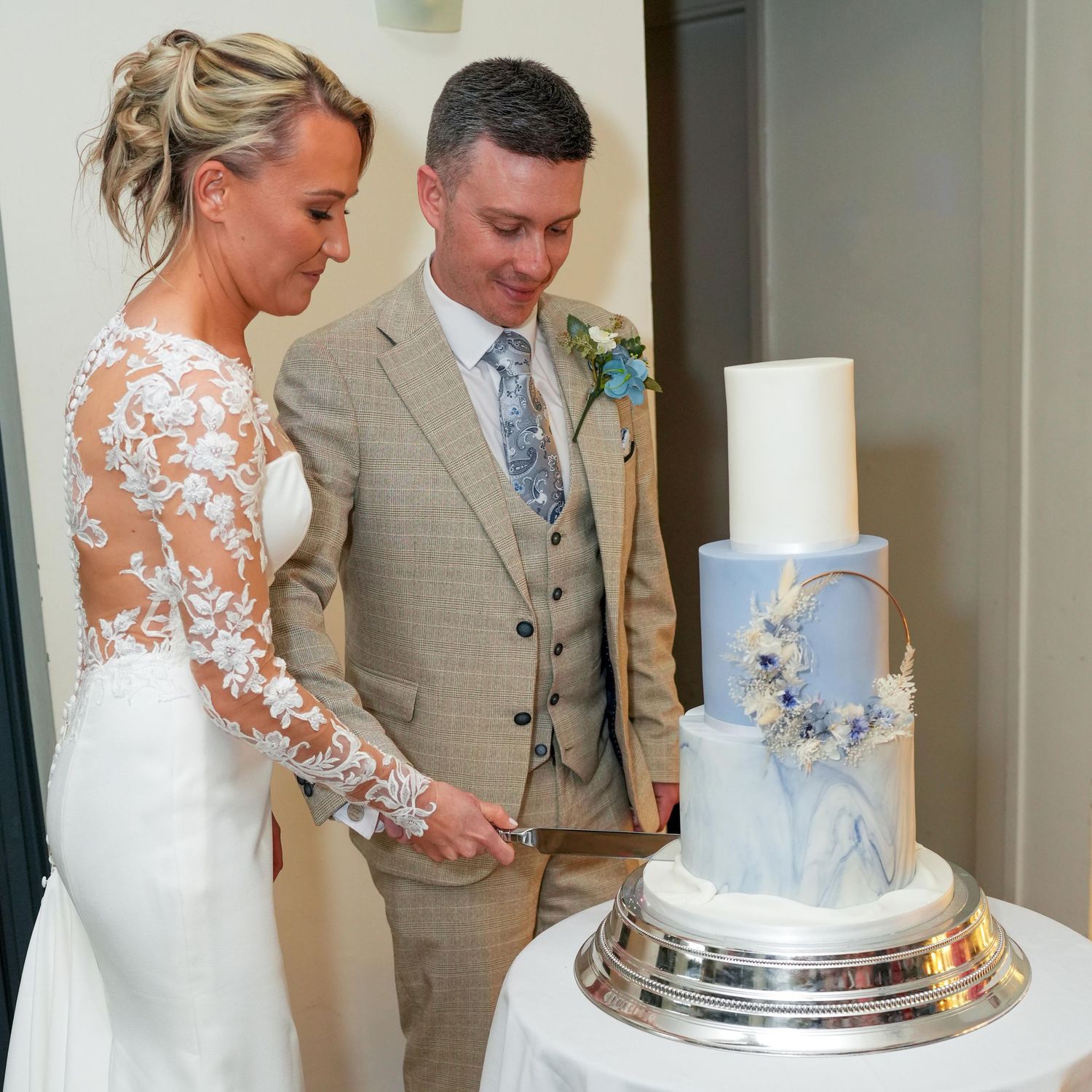 Bride and groom cutting a elegant three-tier white marble wedding cake decorated with blue flowers and gold hoop.
