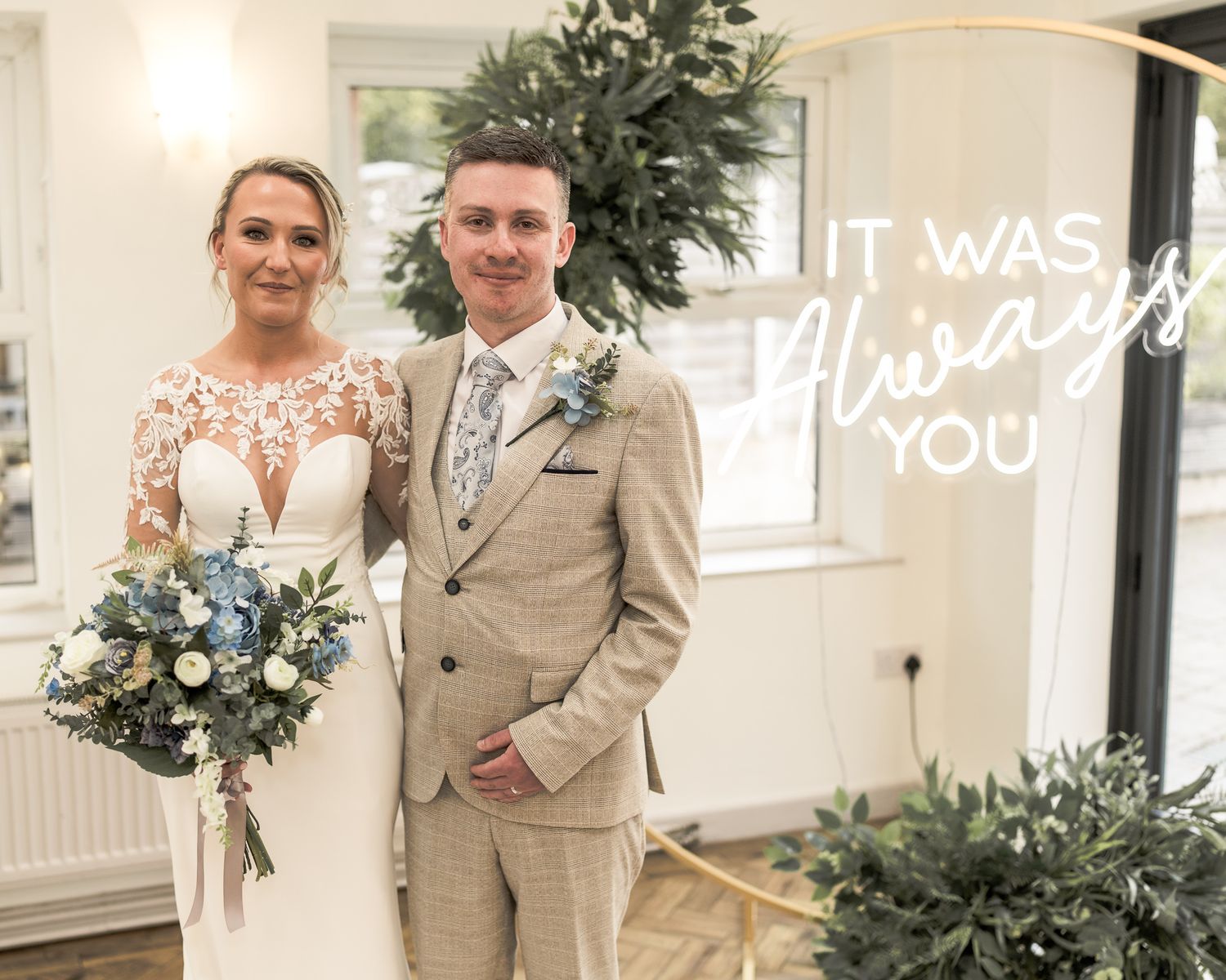 Bride and groom pose together at wedding ceremony with floral arch and neon sign reading It Was Always You.