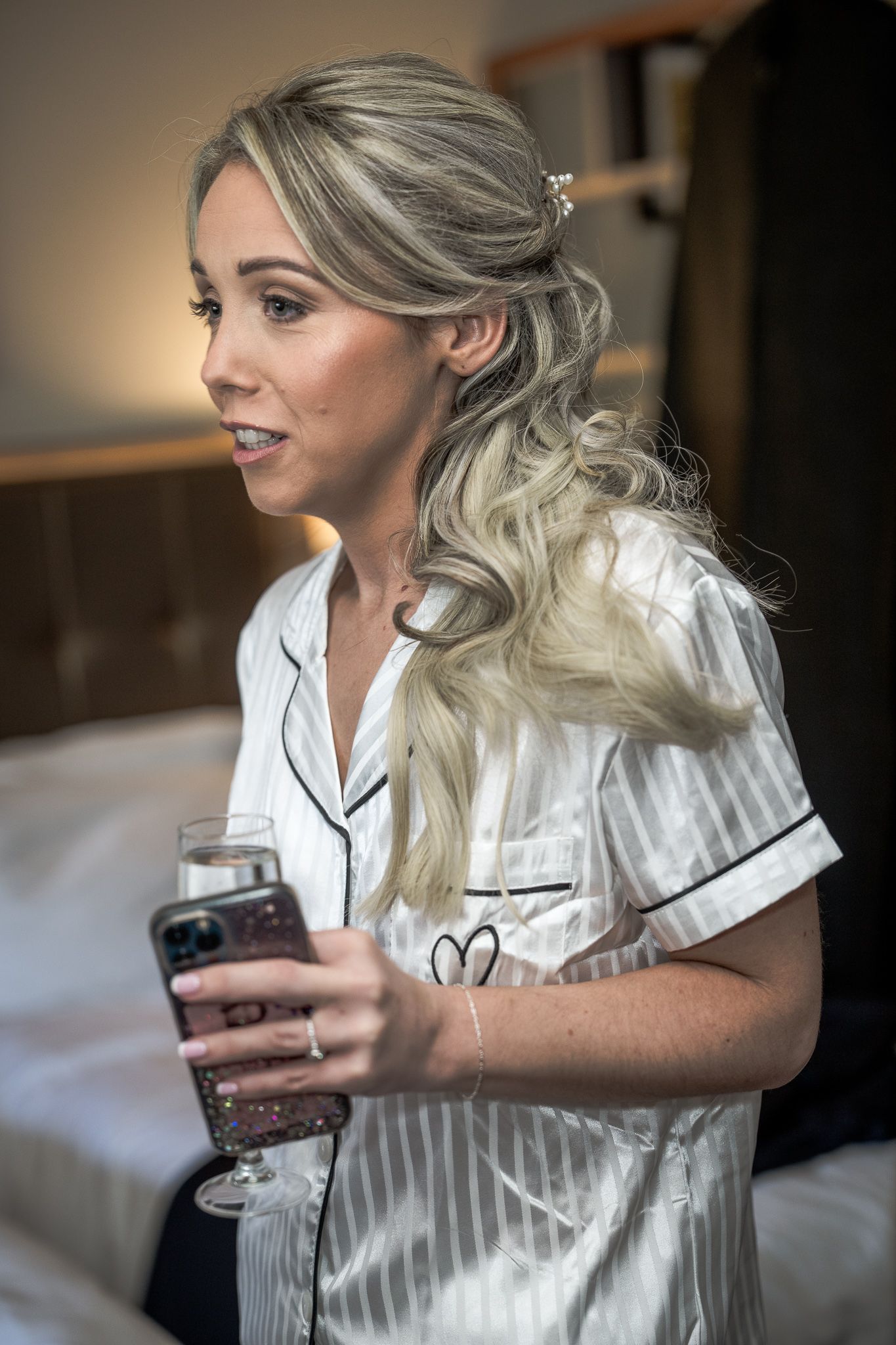 Blonde bride in striped pajamas holding champagne glass and phone while getting ready on wedding day. Dave Martin wedding Photographer NI