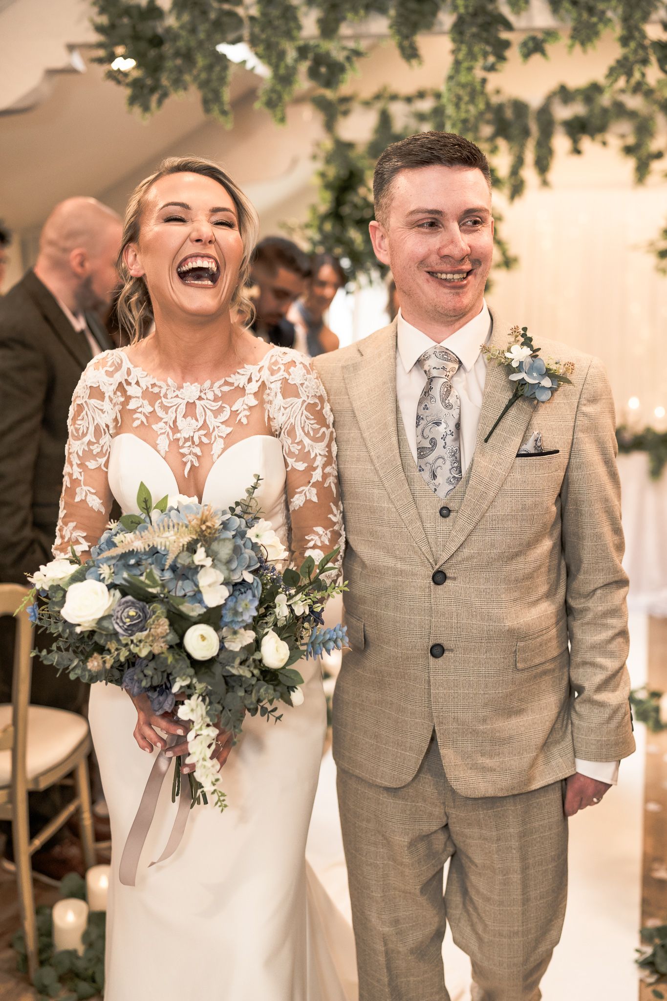Joyful bride in lace gown holding blue and white bouquet walks with groom in tan suit after wedding ceremony. Dave Martin wedding Photograph