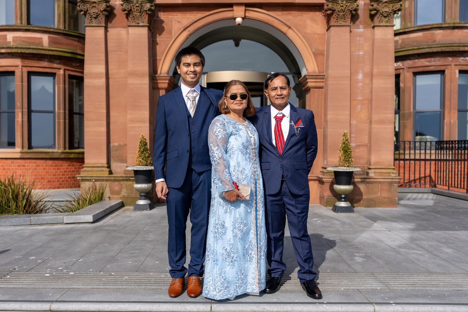 Three elegantly dressed people posing together outside a grand red sandstone building on a sunny day. Slieve Donard Hotel