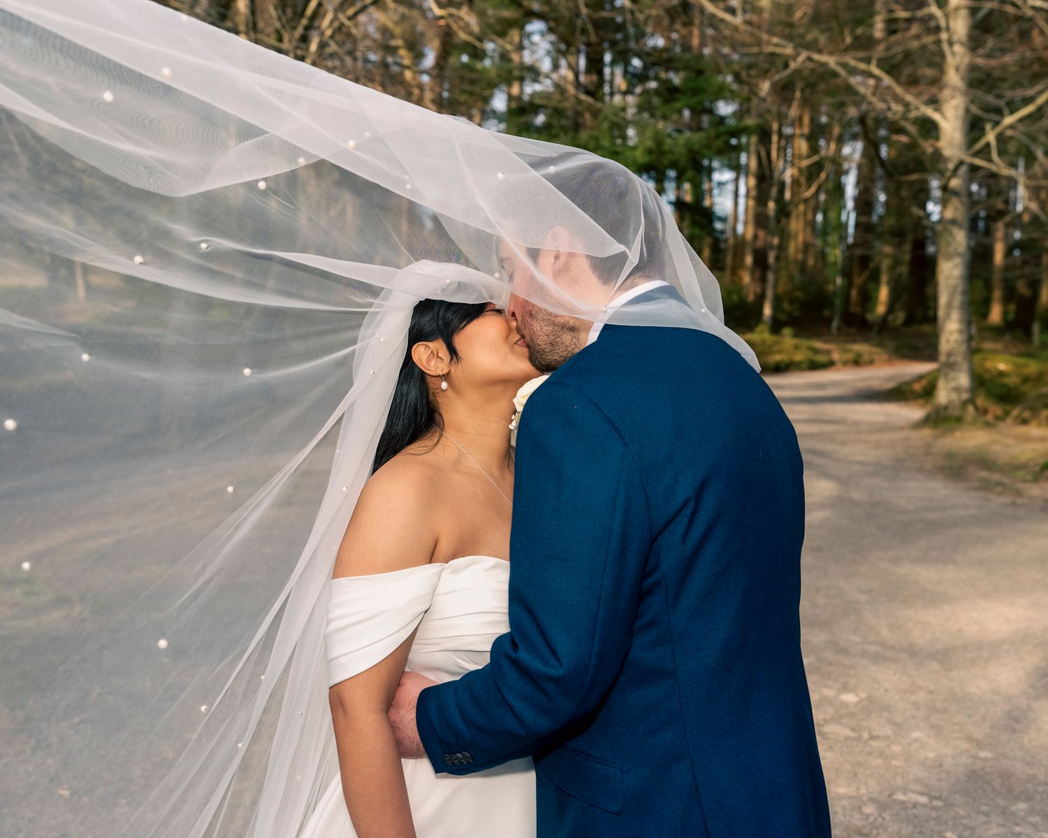 A bride and groom share a romantic kiss under a flowing white veil outdoors, surrounded by tall trees. Dave Martin Photography