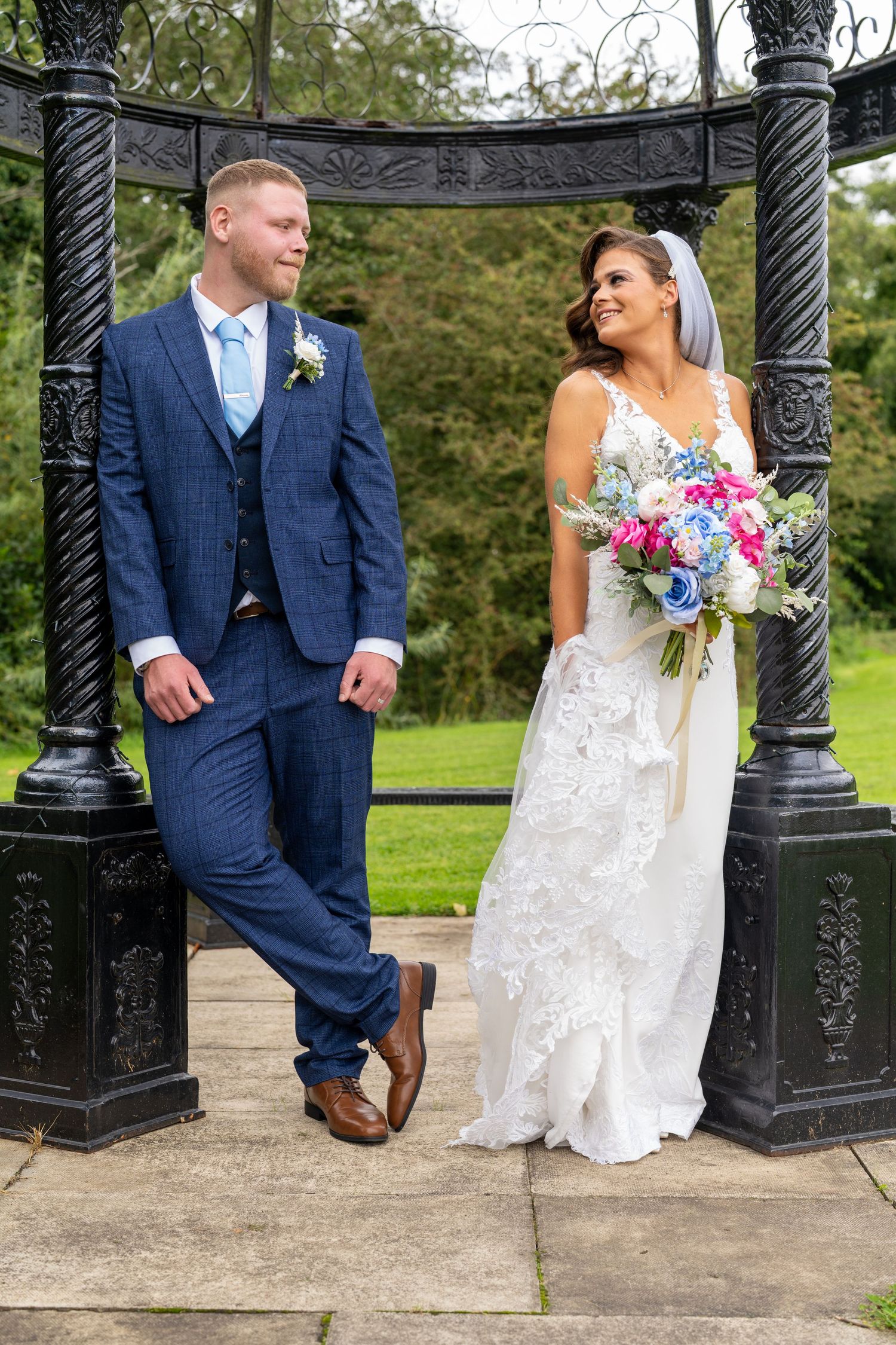 Bride and groom standing under ornate black gazebo, exchanging glances in garden setting. Dave Martin Photographer Northern Ireland