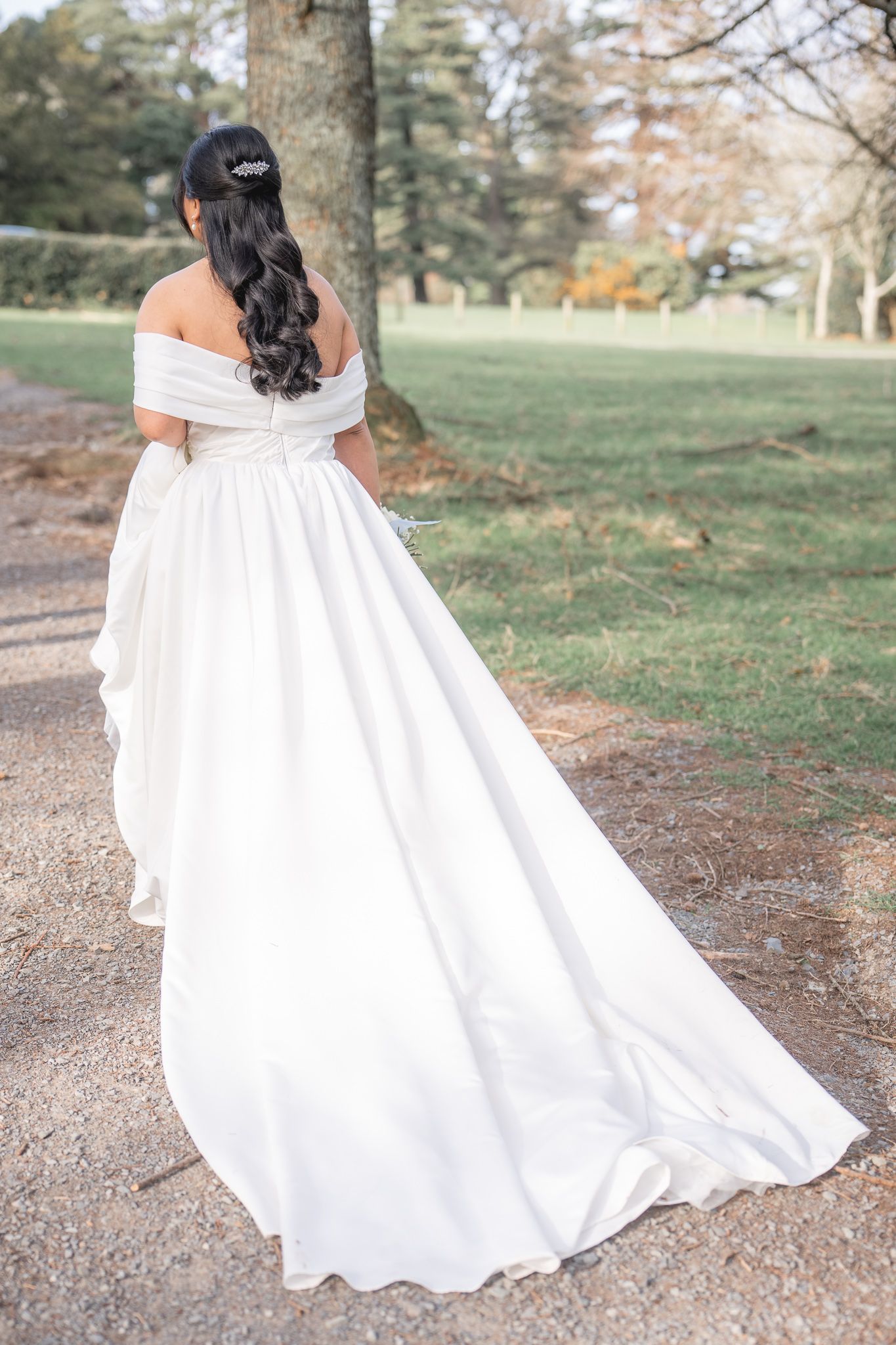Bride in off-shoulder white ball gown with flowing train, dark hair half-up, standing in park. Dave Martin wedding Photographer NI