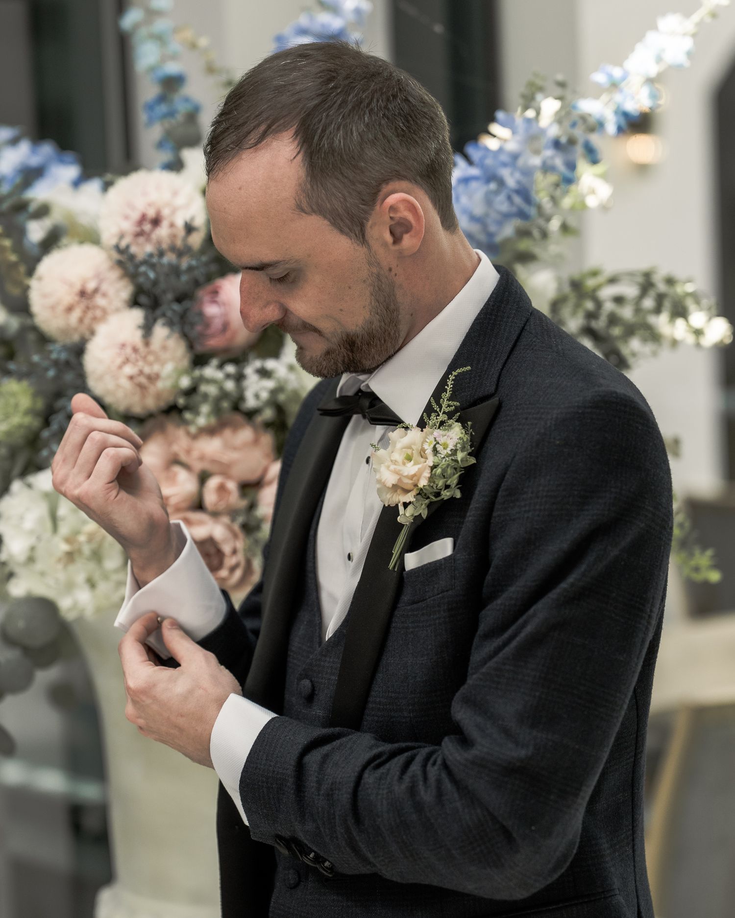 Groom in dark tuxedo adjusting cufflinks with floral boutonniere, elegant wedding flowers in background. Dave Martin wedding Photographer NI
