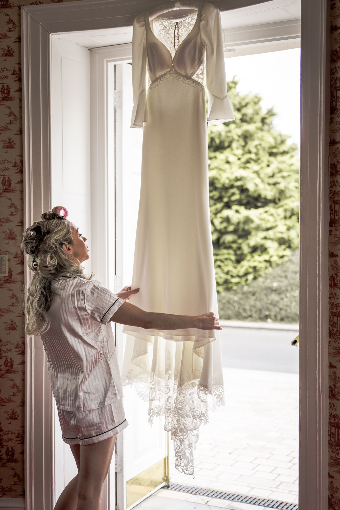 Bride in pajamas with hair curlers admiring her elegant white lace wedding dress hanging in a sunlit doorway.