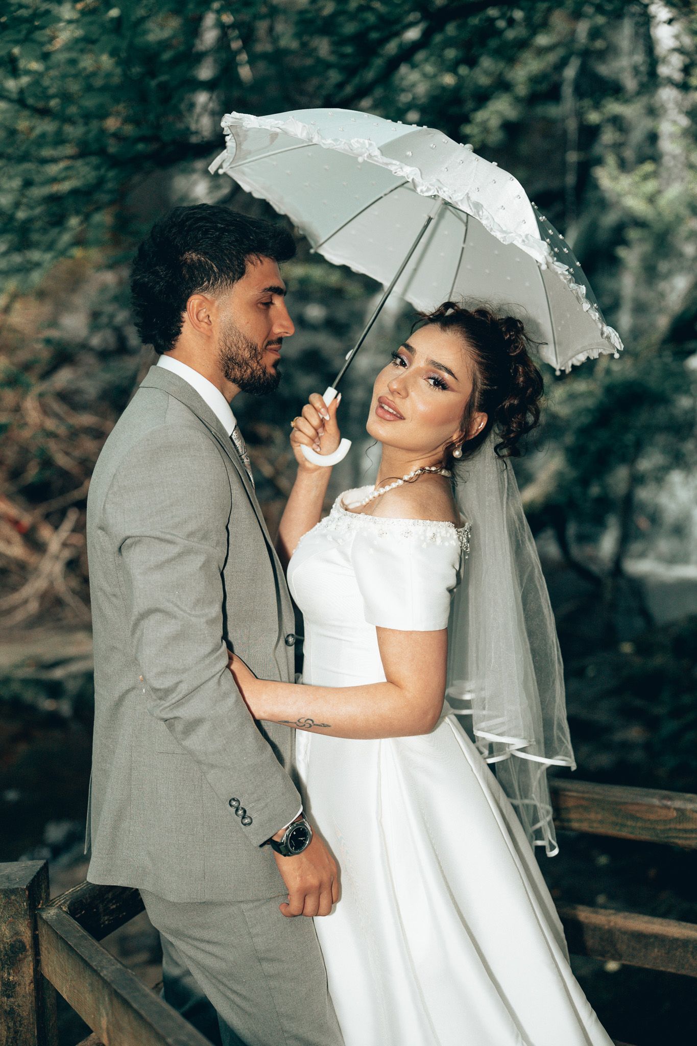 Bride in white gown holds lace umbrella beside groom in gray suit on wooden bridge in lush green forest.
