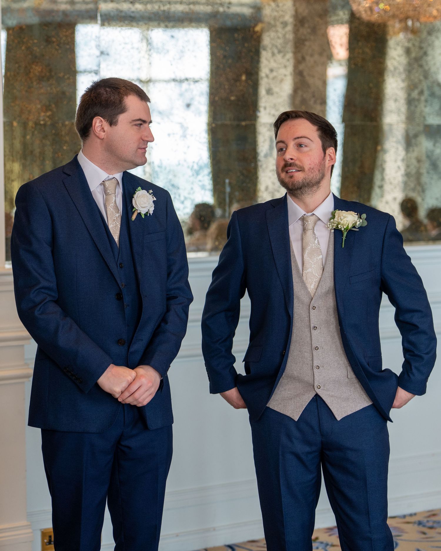 Two groomsmen in navy suits with boutonnieres exchanging glances in elegant mirrored room. Dave Martin Photographer Northern Ireland