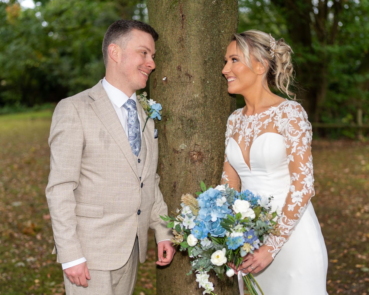 Bride and groom smiling at each other by a tree, bride holding blue and white bouquet in lace wedding dress.