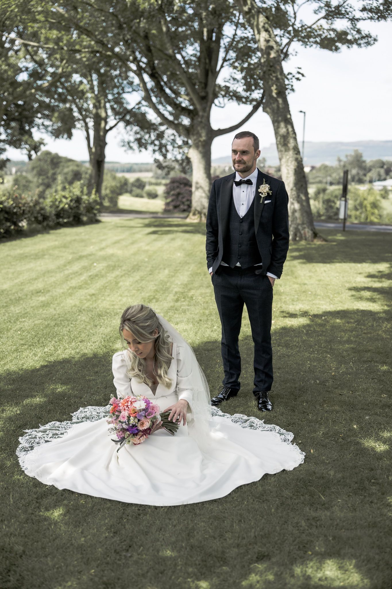 Bride in white gown sits on grass holding bouquet while groom in black tuxedo stands behind her in lush garden.