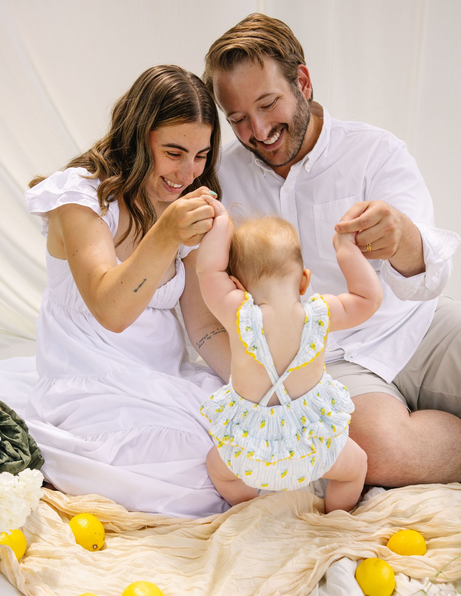Smiling parents holding baby's hands as she sits in a lemon-print romper on a blanket.