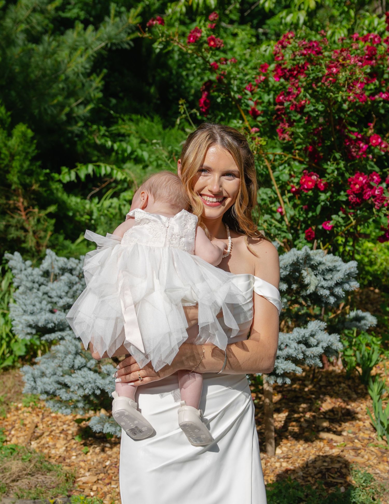 Smiling bride in white off-shoulder gown holds infant dressed in white tutu in sunny garden.