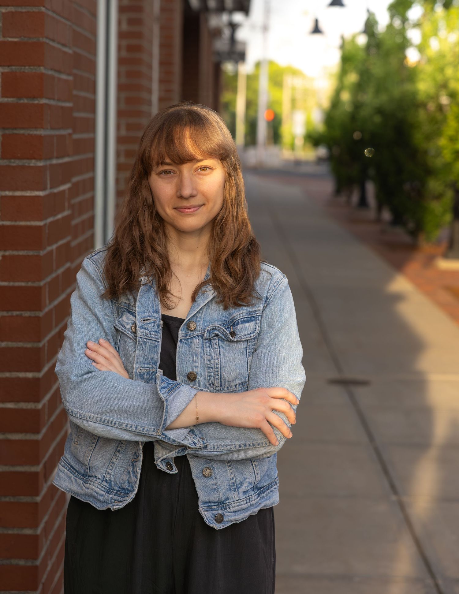 Young woman in denim jacket stands arms crossed on urban brick sidewalk.