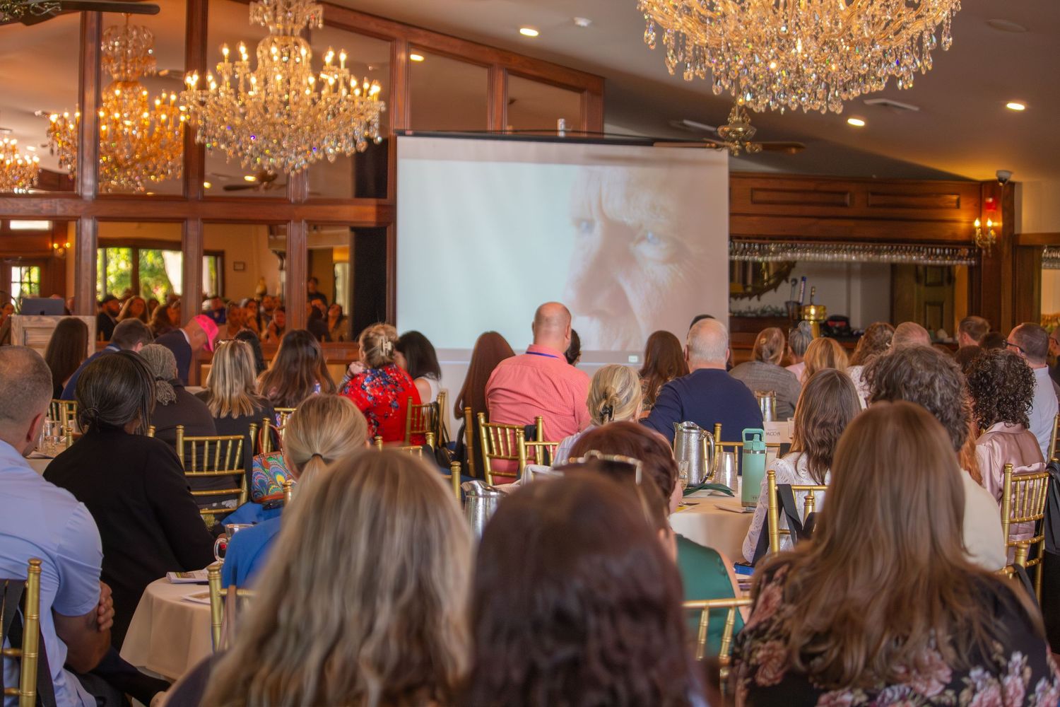 Large audience seated at banquet tables watching a presentation screen in an elegant chandelier ballroom.