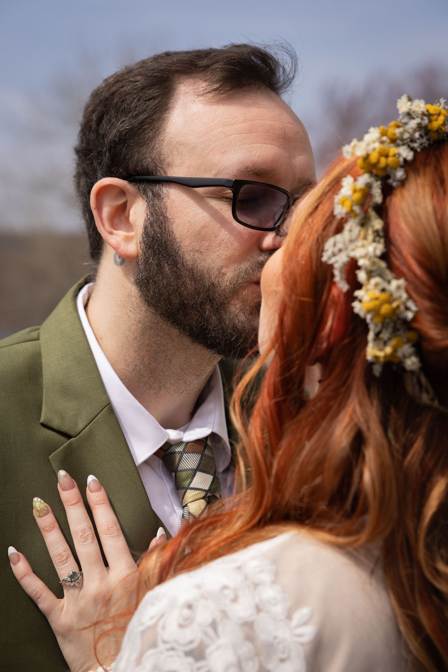 Bearded groom in green suit kissing redhead bride wearing dried flower crown outdoors.