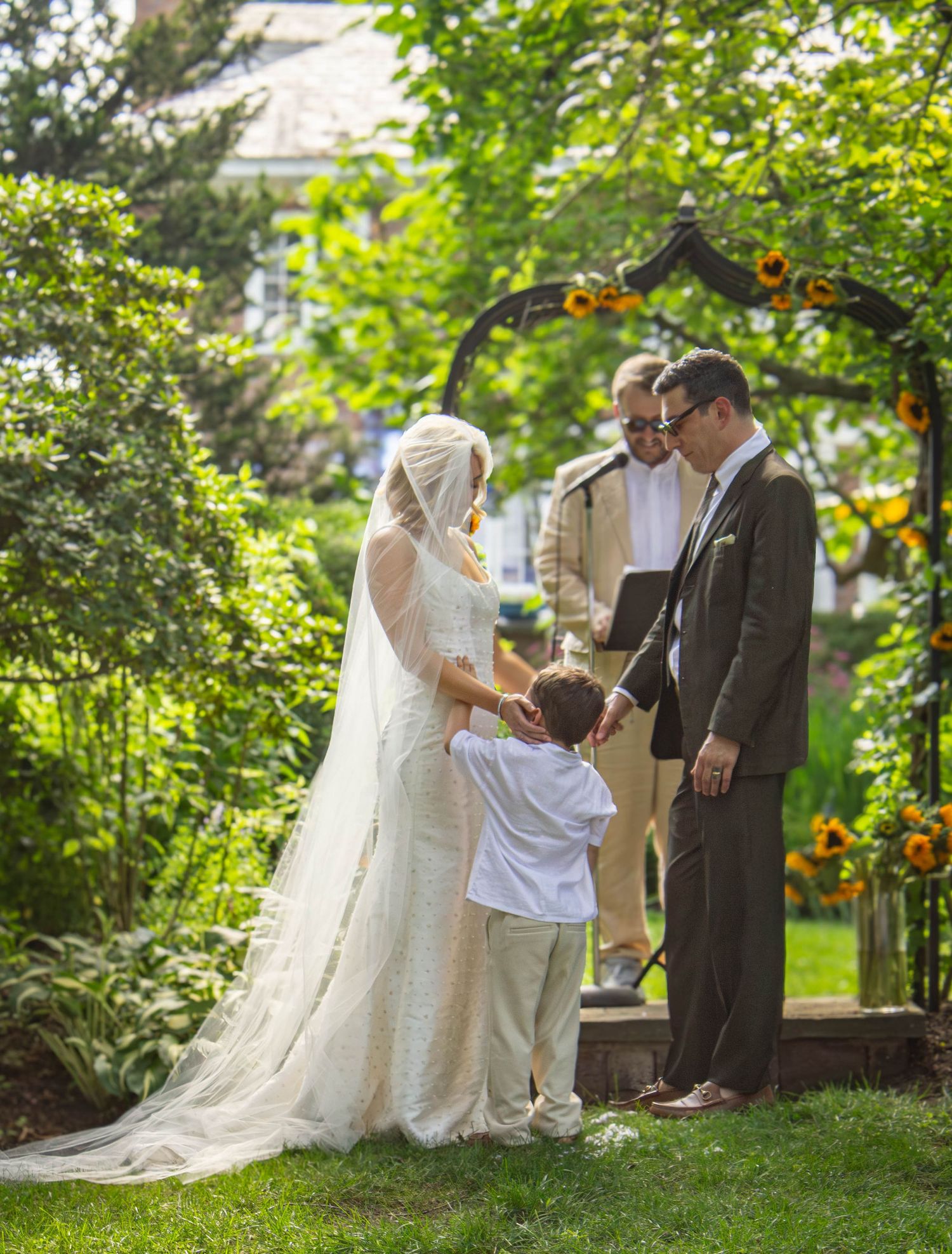 Bride, groom, and child exchange vows under sunflower arch with officiant in lush garden ceremony.