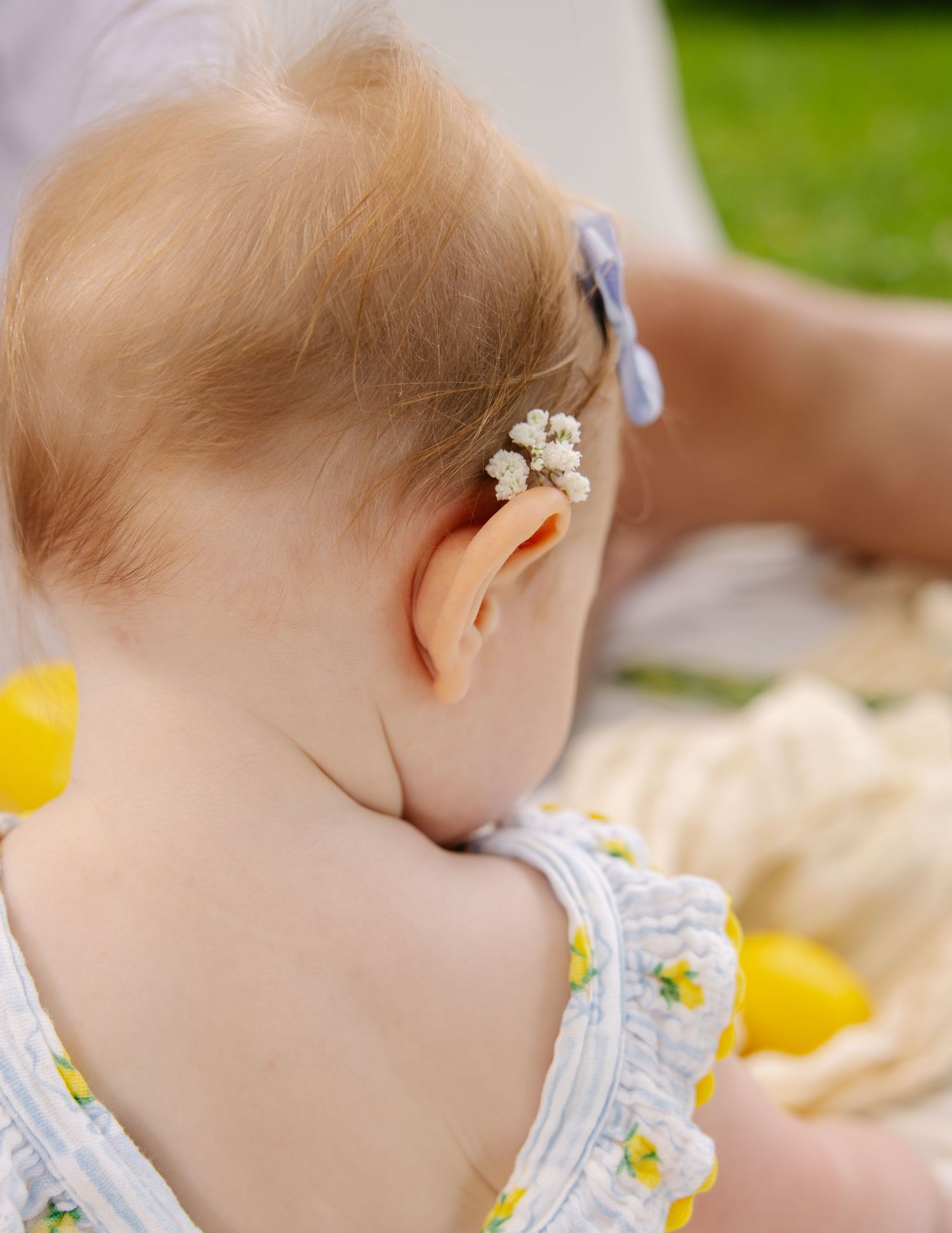 Close-up of baby's head with a floral clip wearing a lemon-print dress outdoors.