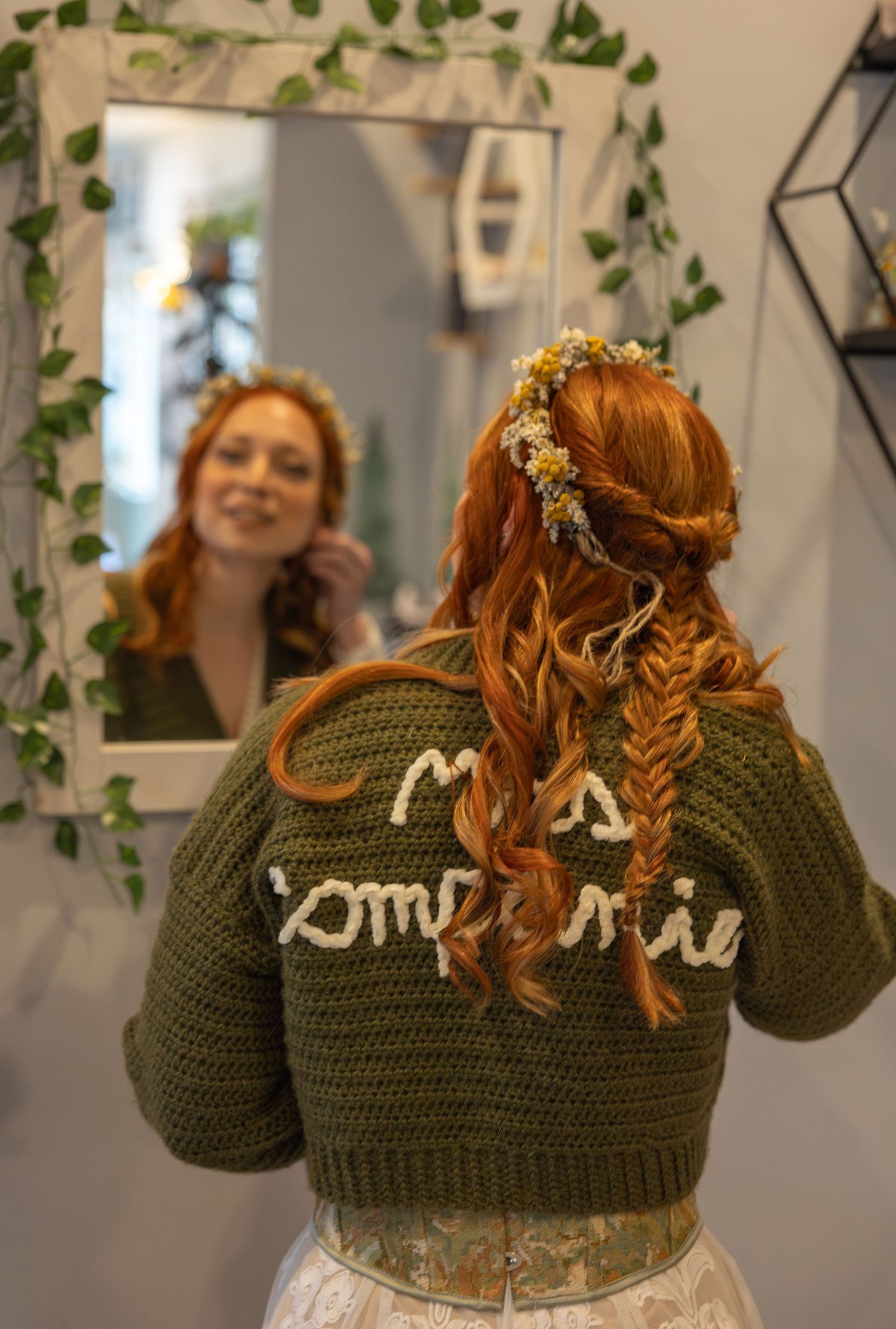 Redheaded bride in green crochet cardigan smiles at mirror reflection with floral crown hairstyle.