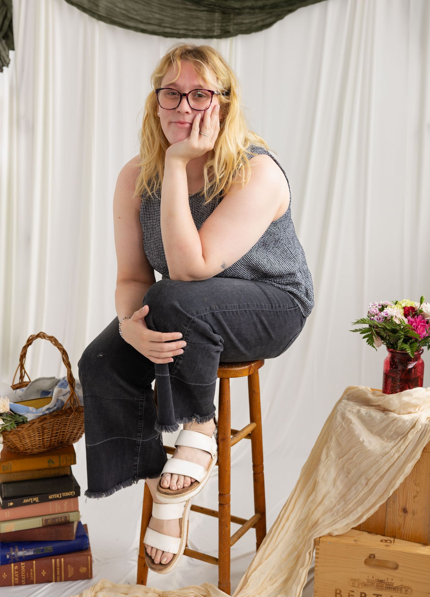Woman with glasses sits on wooden stool in studio portrait setting with flowers and books.