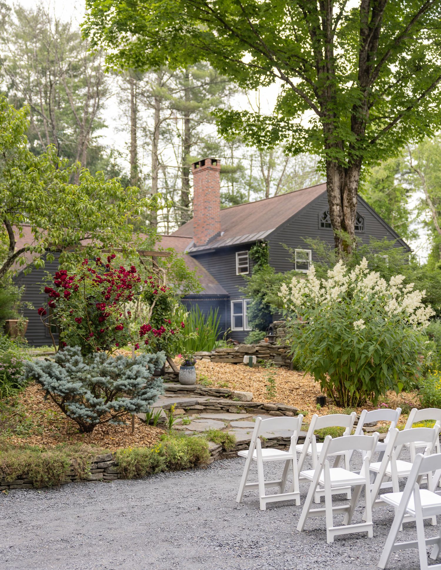 White chairs arranged for outdoor wedding ceremony beside charming dark cottage surrounded by lush gardens.