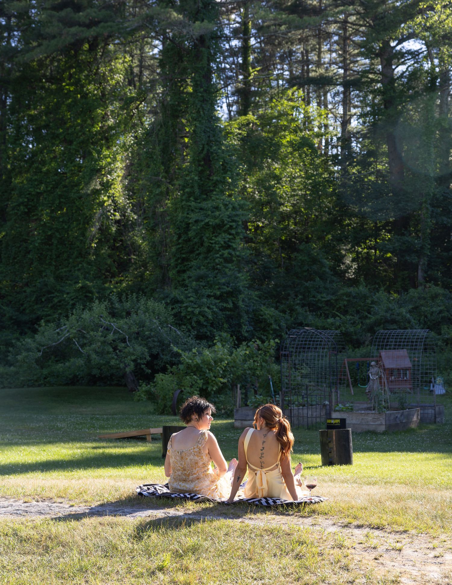 Two young girls in matching gold dresses sit together on a sunny lawn surrounded by trees.
