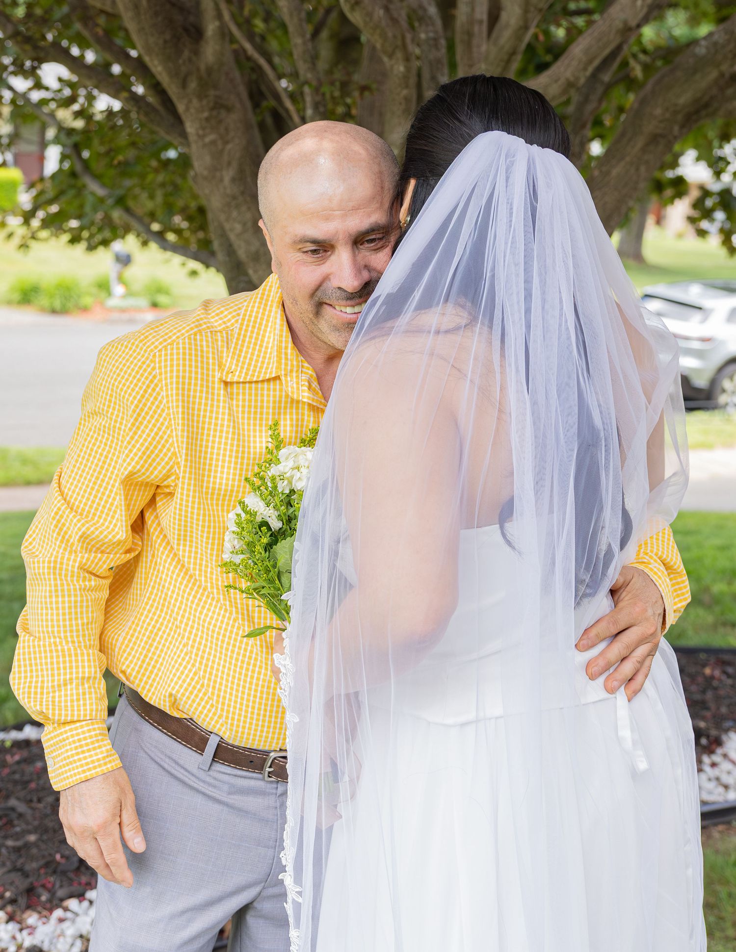 Emotional father hugging bride in white wedding dress and veil outdoors before ceremony.